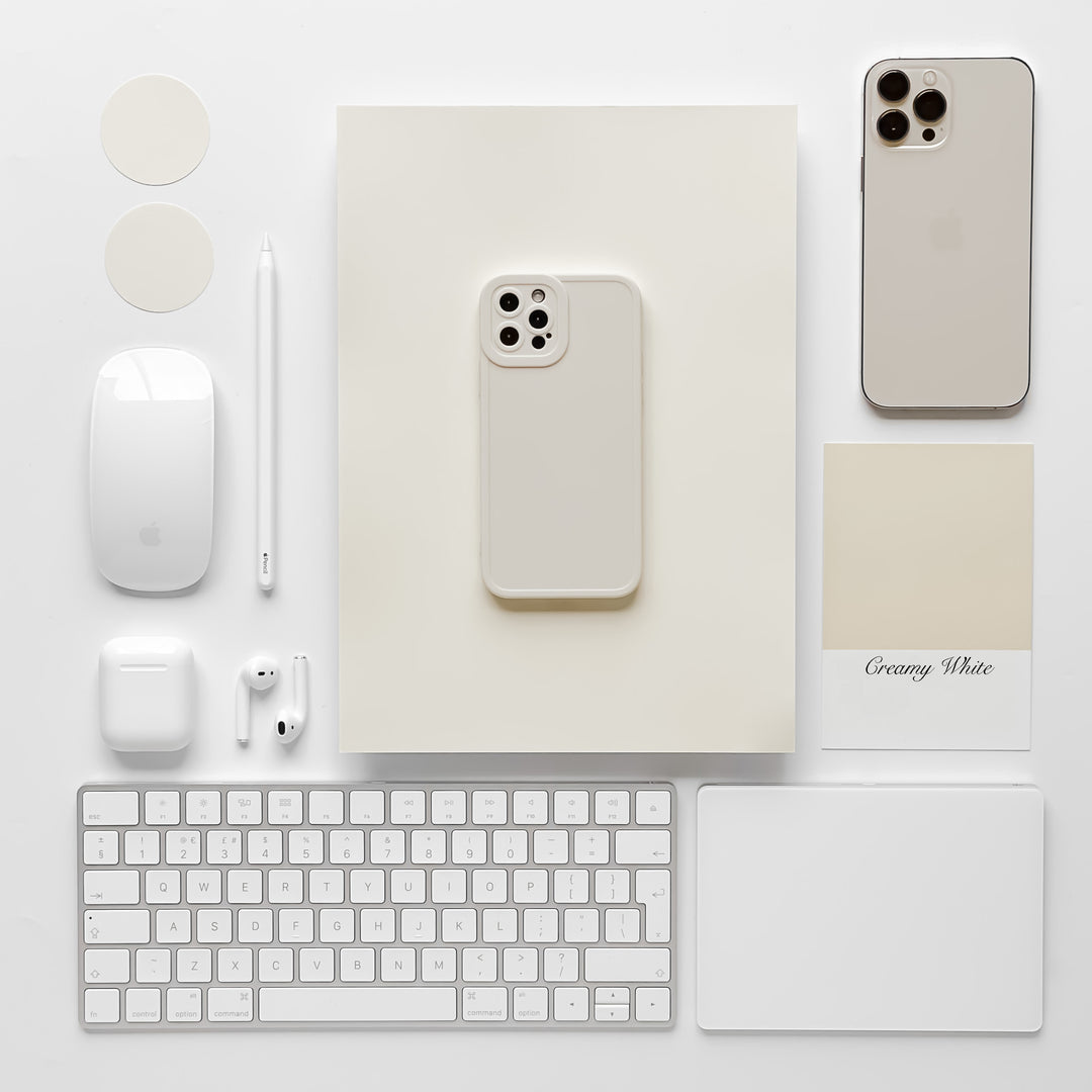 A flat lay image showcasing a collection of white tech gadgets and accessories neatly arranged on a white background. It features an iPhone with the NAKD Phone Case - Creamy White by Coconut Lane, a wireless mouse, keyboard, AirPods, Apple Pencil, round stickers, a mouse pad, and a paint swatch labeled "Creamy White.