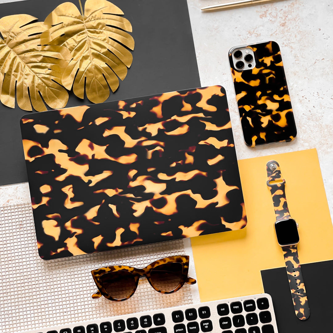 Flat lay photo showcasing a collection of coordinated tech and fashion accessories adorned with a tortoiseshell pattern. Included in the ensemble are a laptop, smartphone case, Coconut Lane's Chic Tortoiseshell Apple Watch Strap with matching band, sunglasses, keyboard, and golden monstera leaves displayed on a mixed black and white surface.