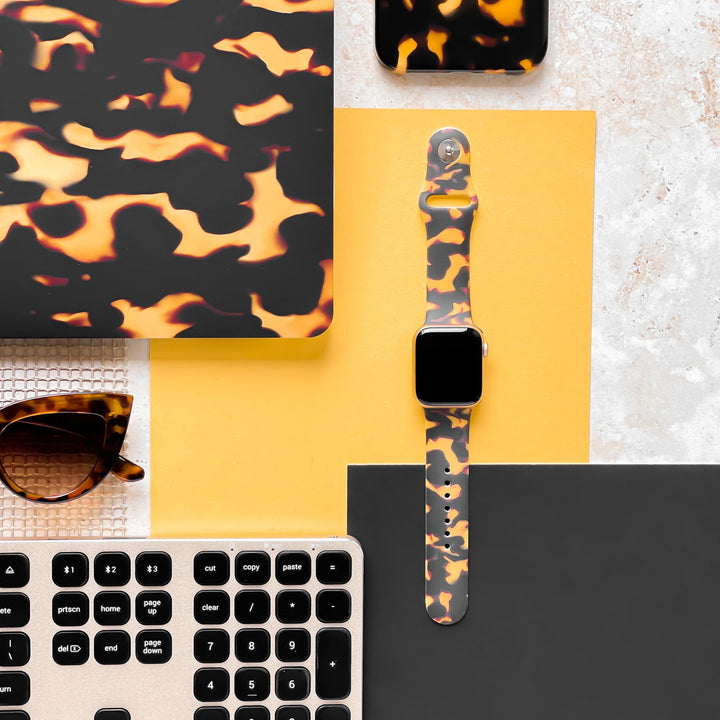 A flat lay image showcasing a matching camouflage-patterned set on a vibrant yellow and gray background. Items included are a laptop sleeve, smartphone case, Chic Tortoiseshell Apple Watch Strap by Coconut Lane, and sunglasses. Adjacent to the ensemble is a laptop keyboard positioned at the bottom edge of the frame.