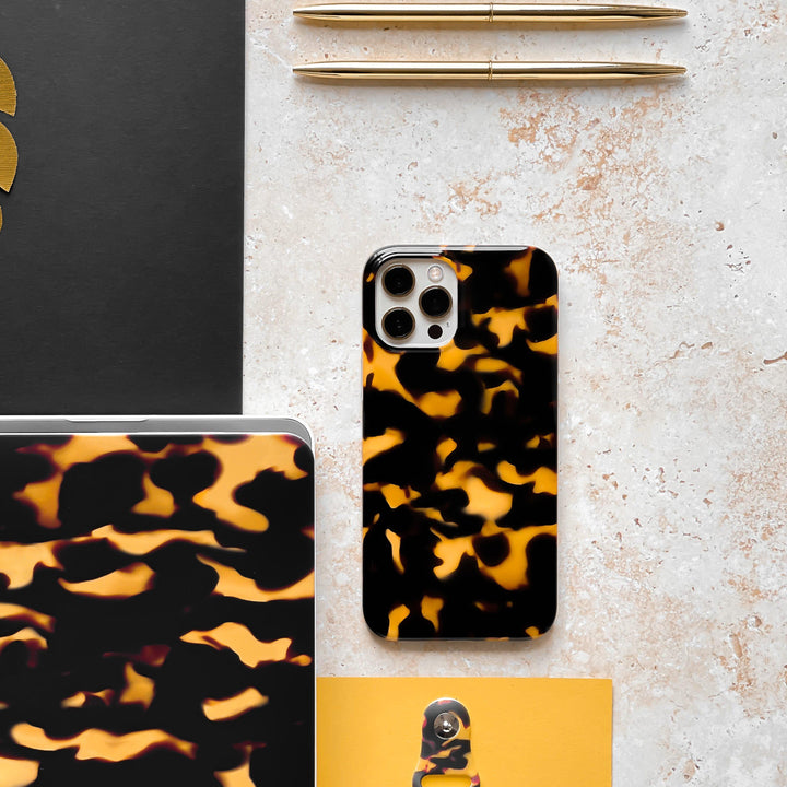 This image showcases a flat lay of office supplies on a beige marble surface, featuring two gold pens, a closed laptop, a notebook, and a clipboard. The focus is on a smartphone adorned with the Chic Tortoiseshell Phone Case by Coconut Lane, perfectly matching the laptop cover.