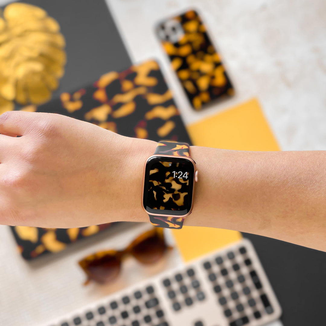 A person wearing Coconut Lane's Chic Tortoiseshell Apple Watch Strap, which displays the time "1:24." In the background, there's a matching patterned smartphone case, laptop cover, and sunglasses, along with a keyboard and a yellow notebook.