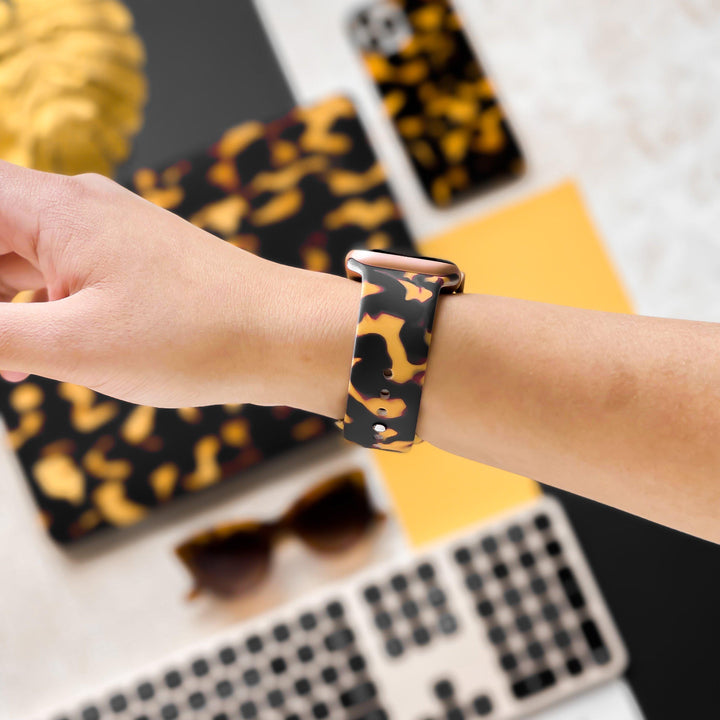 A hand sporting a Chic Tortoiseshell Apple Watch Strap by Coconut Lane is seen. In the background, objects matching the camouflage theme, such as a laptop cover, phone case, and a pair of sunglasses are visible. A keyboard and some yellow and black items can also be seen.