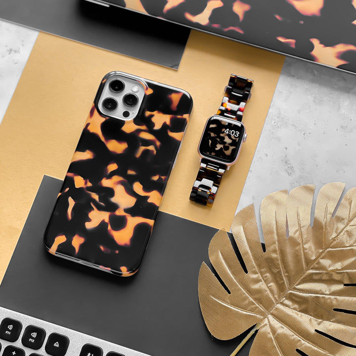 A smartphone encased in Coconut Lane's Chic Tortoiseshell Phone Case rests on a black and gold surface beside a matching smartwatch showing 4:03. The scene is complemented by a silver laptop, part of a keyboard, and an ornamental golden leaf.