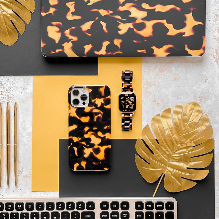 A flat lay of an organized, stylish workspace featuring the Chic Tortoiseshell Phone Case from Coconut Lane made from soft flexible plastic, a matching laptop sleeve, and a watch on a yellow and black background. Golden pens and metallic leaves add an elegant touch, alongside a sleek black keyboard on a marble surface.