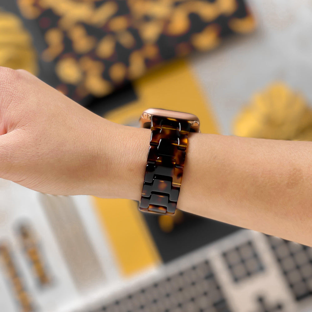 A close-up of a left wrist adorned with the Coconut Lane Luxe Tortoiseshell Apple Watch Strap, featuring a rose gold watch face. In the background, blurred objects display a mix of yellow, black, and gold hues. The adjustable strap ensures the perfect fit for various Apple Watch models.