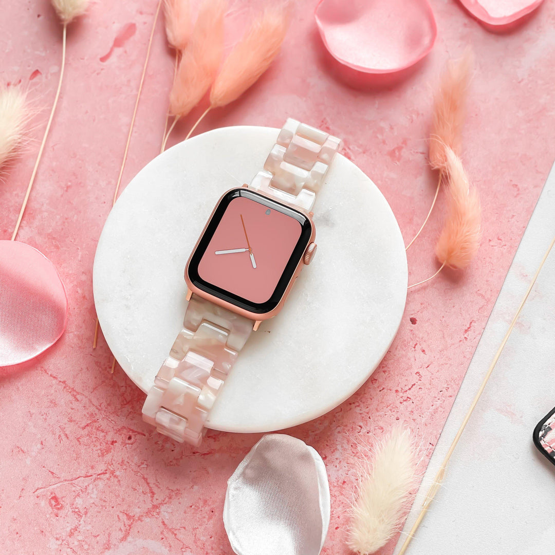 The Luxe Rose Apple Watch Strap from Coconut Lane, featuring a pink face and a marble-patterned, adjustable band, is elegantly displayed on a round white pedestal. Pink rose petals and fluffy stems are artistically scattered around, set against a pink textured background. The overall aesthetic is soft and elegant.