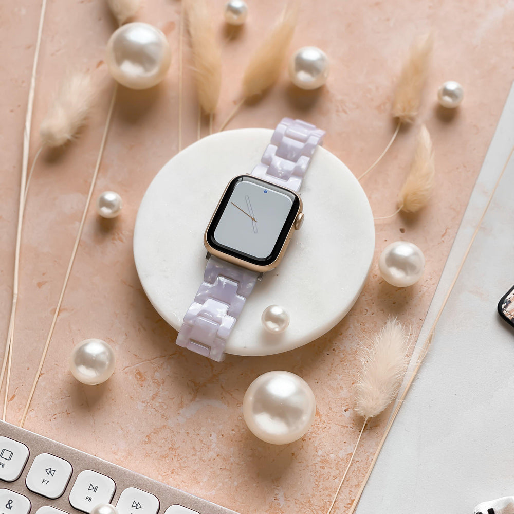 A smartwatch adorned with the Luxe Pearl Apple Watch Strap by Coconut Lane is displayed on a circular white stand. Scattered pearls and delicate dried plants surround it, set against a light pink marble surface. The corner of a white keyboard peeks into the frame from the bottom left corner.