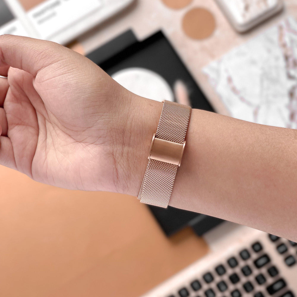 Close-up of a person's wrist adorned with the Coconut Lane Milanese Apple Watch Strap in Rose Gold. The background reveals a blurred desk featuring a keyboard, notebooks, and various stationery items, all set against a warm and neutral color scheme.