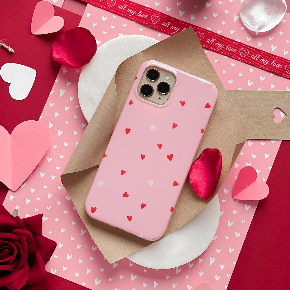A Coconut Lane Cute Hearts Phone Case, adorned with small red and white hearts, is placed on a brown envelope. Surrounding it are red and white paper hearts and rose petals. The background highlights a red and pink color scheme with a ribbon that reads "all my love.