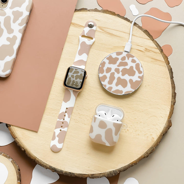 A wooden surface displays a smartwatch with a Nude Cow Apple Watch Strap by Coconut Lane, a wireless charging pad with matching vibes, and wireless earbuds in a silicone cow-print case. Nearby, there is a brown rectangular object and portions of similarly patterned items.