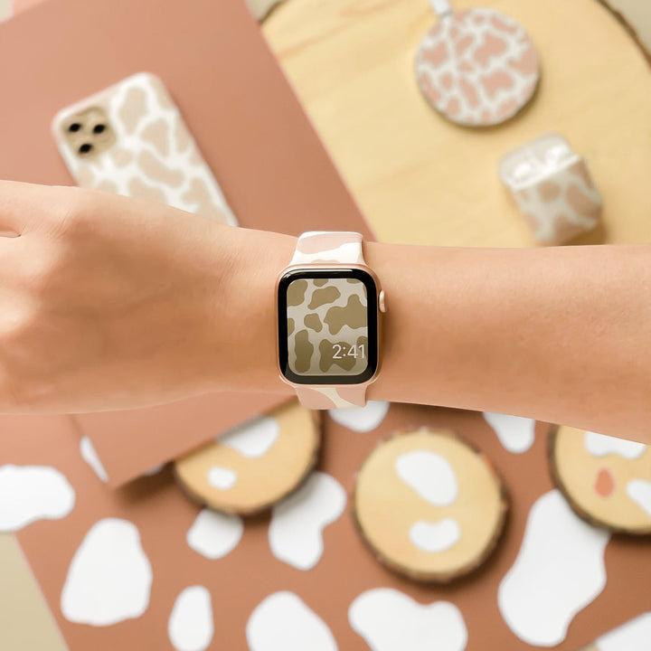 A wrist dons a Coconut Lane smartwatch with a Nude Cow Apple Watch Strap displaying the time "2:41" and sporting a beige and white cow print design. Surrounding items exude matching vibes, including a phone case, reusable tote bag, notebook, and accessories, all featuring the same chic cow print pattern.