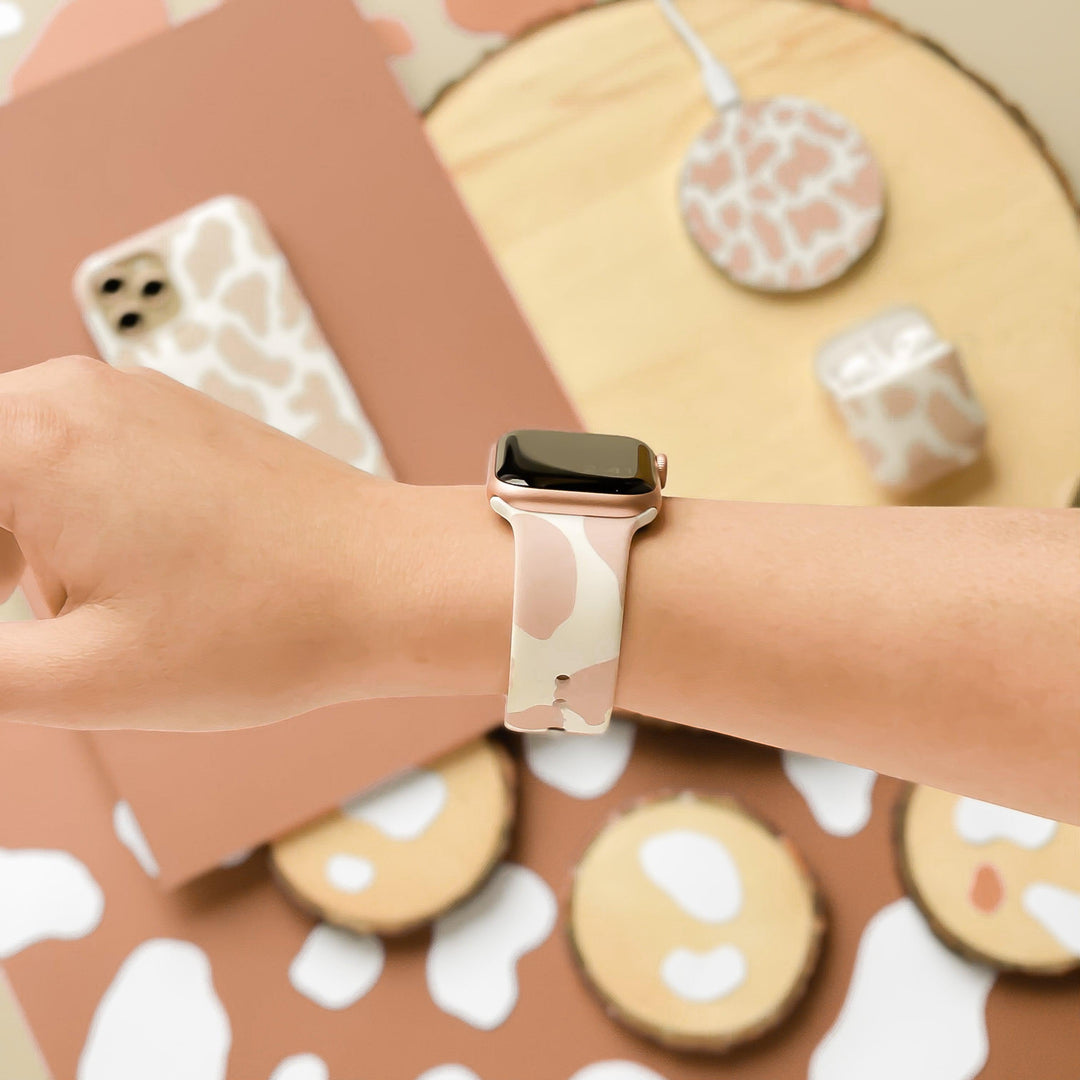 A person wearing a Coconut Lane Nude Cow Apple Watch Strap with a beige and white patterned band. The background exudes matching vibes, featuring various tech accessories like a phone case, wireless charger, and AirPods case in the same pattern on a light wooden surface. The silicone finish adds a modern touch.