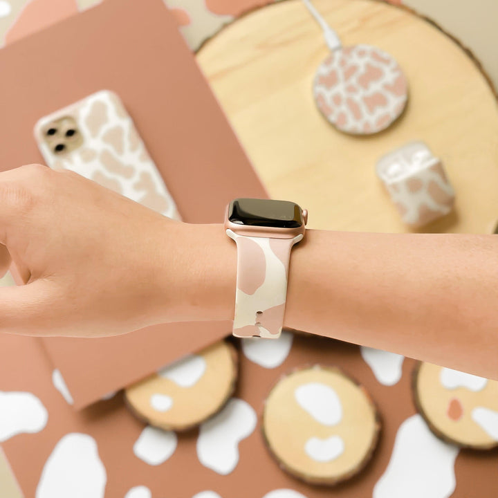 A person wearing a Coconut Lane Nude Cow Apple Watch Strap with a beige and white patterned band. The background exudes matching vibes, featuring various tech accessories like a phone case, wireless charger, and AirPods case in the same pattern on a light wooden surface. The silicone finish adds a modern touch.