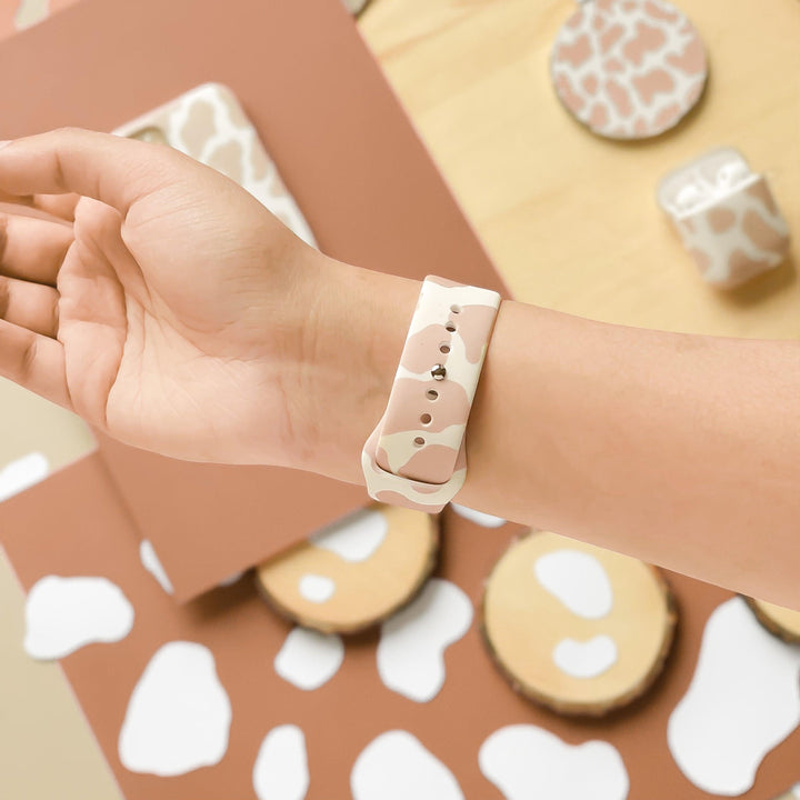 A person's wrist is shown wearing the Nude Cow Apple Watch Strap from Coconut Lane, featuring a beige and light brown cow pattern on silicone. The background includes various items such as phone cases and circular objects, all showcasing the same matching vibes cow pattern.