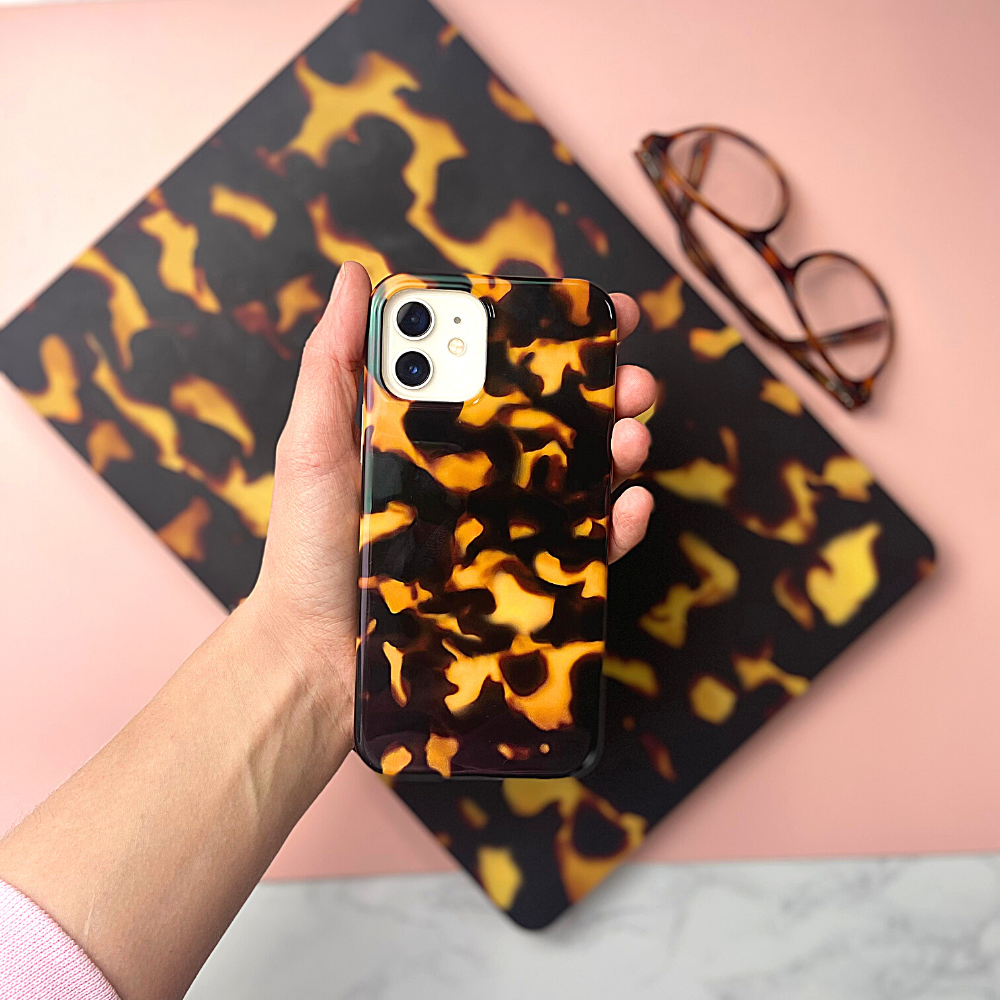 A hand holds Coconut Lane's Chic Tortoiseshell Phone Case, crafted from soft flexible plastic. In the background, there's a matching tortoiseshell-patterned item and a pair of eyeglasses resting on a pink surface. The scene is set on a light-colored marble table, all enhanced by IMD Print Technology for lasting beauty.