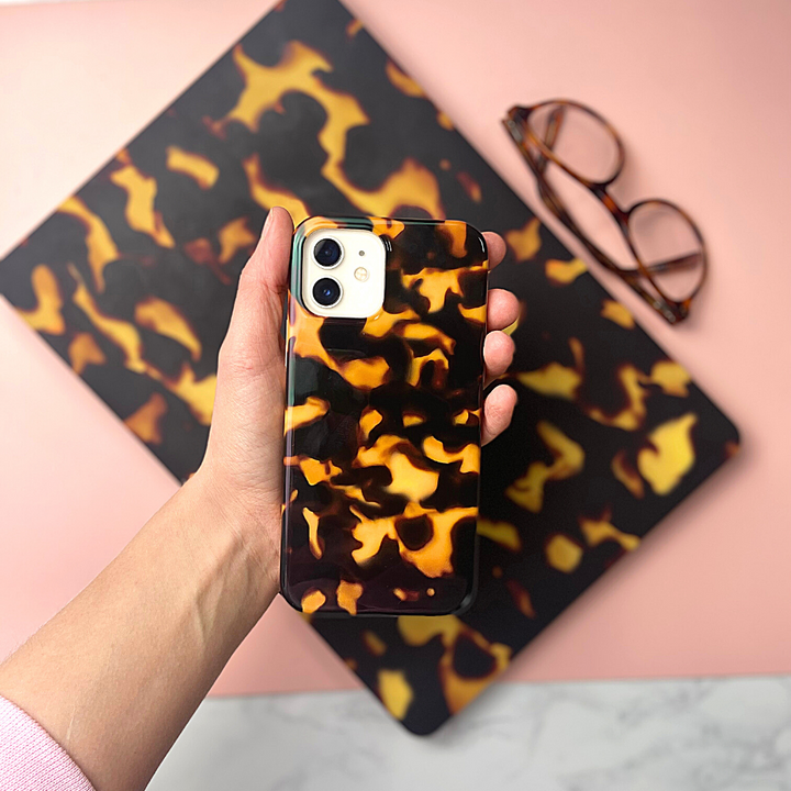 A hand holds Coconut Lane's Chic Tortoiseshell Phone Case, crafted from soft flexible plastic. In the background, there's a matching tortoiseshell-patterned item and a pair of eyeglasses resting on a pink surface. The scene is set on a light-colored marble table, all enhanced by IMD Print Technology for lasting beauty.