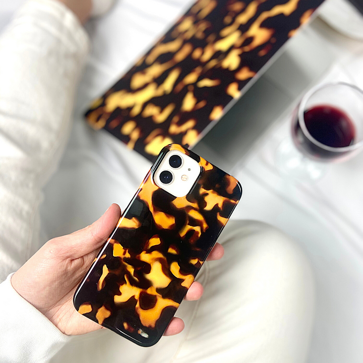 A person holding the Chic Tortoiseshell Phone Case from Coconut Lane made with IMD Print Technology. The background includes a matching tortoiseshell-patterned laptop cover, a glass of red wine, and part of the person's white pants, suggesting a cozy setting.