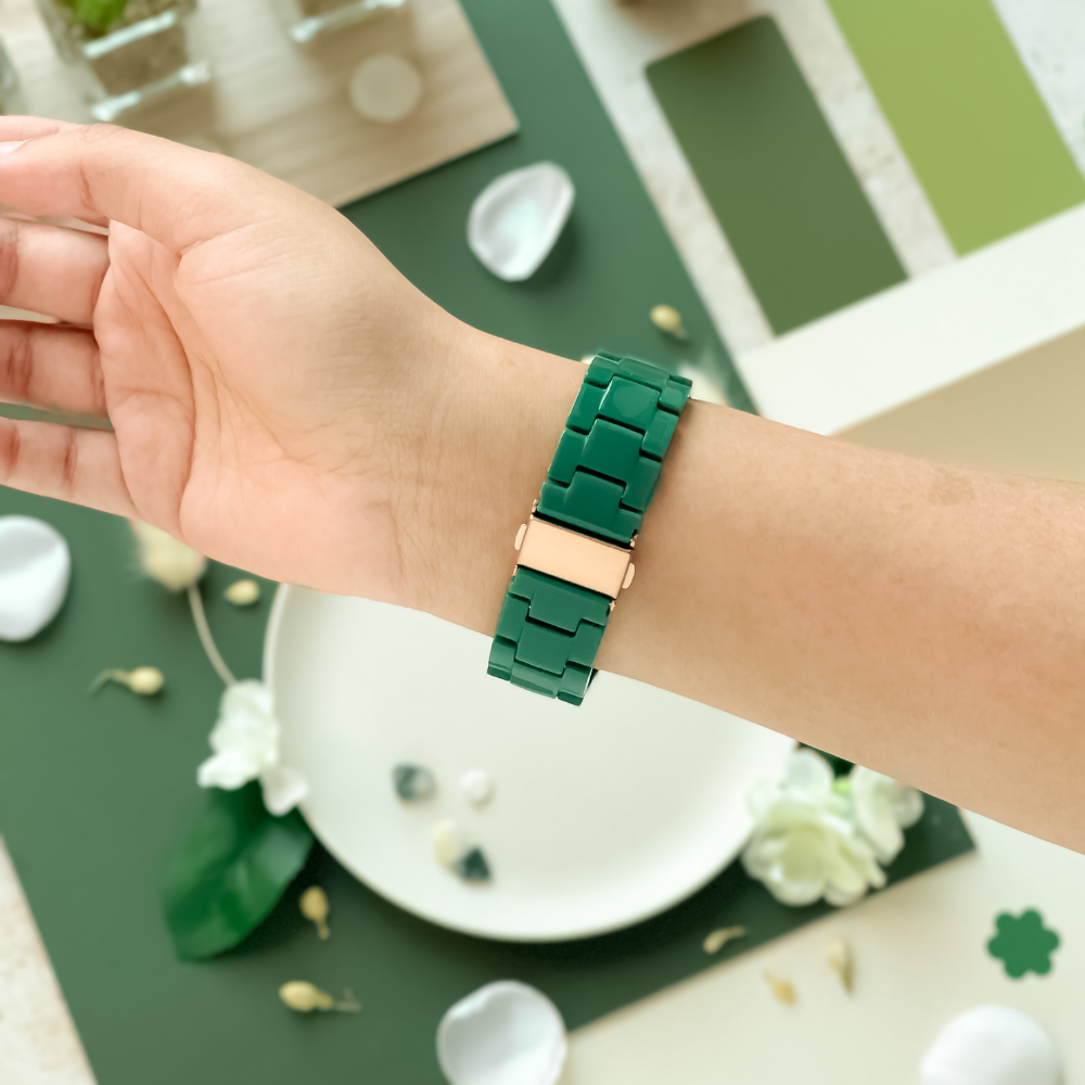 A person's wrist is shown wearing a Coconut Lane Luxe Green Apple Watch Strap with a gold clasp. The background includes various green and white objects, such as flowers, petals, and geometric shapes, artfully arranged on a white and green surface.