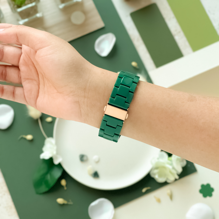 A person's wrist is shown wearing a Coconut Lane Luxe Green Apple Watch Strap with a gold clasp. The background includes various green and white objects, such as flowers, petals, and geometric shapes, artfully arranged on a white and green surface.