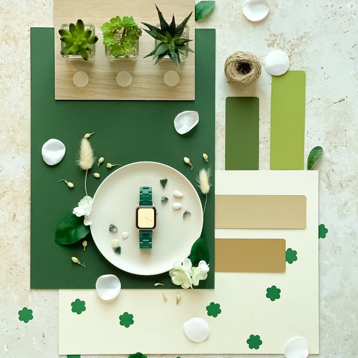 A flat lay photo with a green-themed aesthetic featuring the Luxe Green Apple Watch Strap by Coconut Lane on a white plate, surrounded by leaves, petals, color swatches, succulents in glass pots, a spool of twine, and decorative elements like small clovers and white flowers.