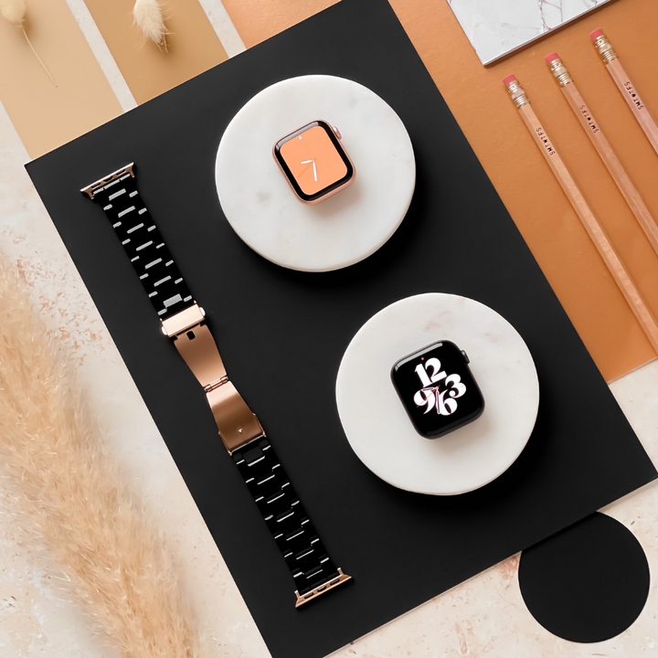 Flat lay image of two smartwatches with black bands and rose gold detailing, displayed on two round white stands placed on a black rectangular surface. A Luxe Black Apple Watch Strap from Coconut Lane, three pencils, beige pampas grass plumes, and neutral-colored papers are also arranged around them.