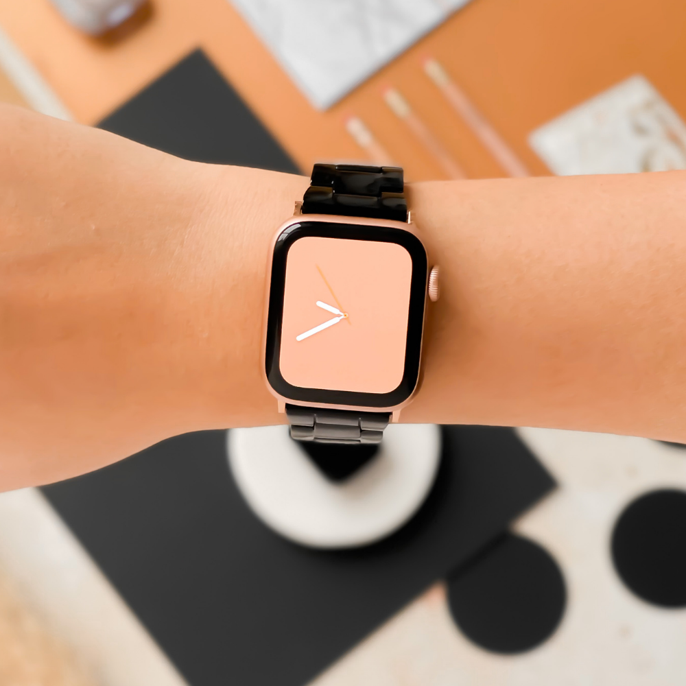 Close-up of a wrist wearing a modern smartwatch with a minimalist design featuring a square face and a blank beige screen displaying simple white clock hands. The ensemble is elevated by the elegant Luxe Black Apple Watch Strap from Coconut Lane. The background includes various blurred objects, hinting at an indoor setting.