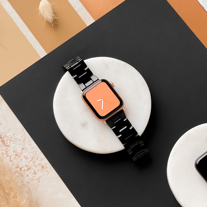 A Coconut Lane Luxe Black Apple Watch Strap is showcased on a round white marble surface. The smartwatch screen features an orange background with a white number seven. The setting includes earthy tones and textures, contributing to a modern aesthetic.