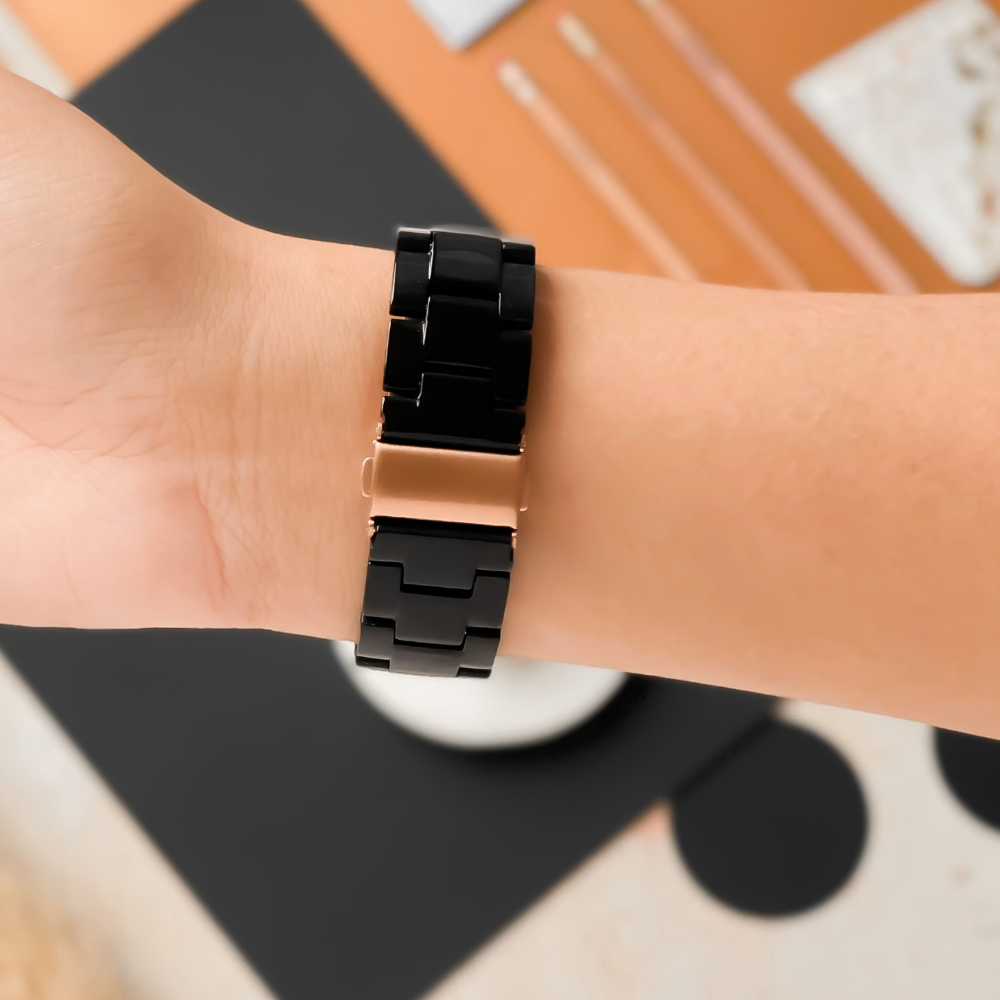 A person is wearing a Coconut Lane Luxe Black Apple Watch Strap, featuring a glossy black link design and a metallic clasp, on their wrist. In the background, there is a minimalist desk setup with pencils and a notepad.
