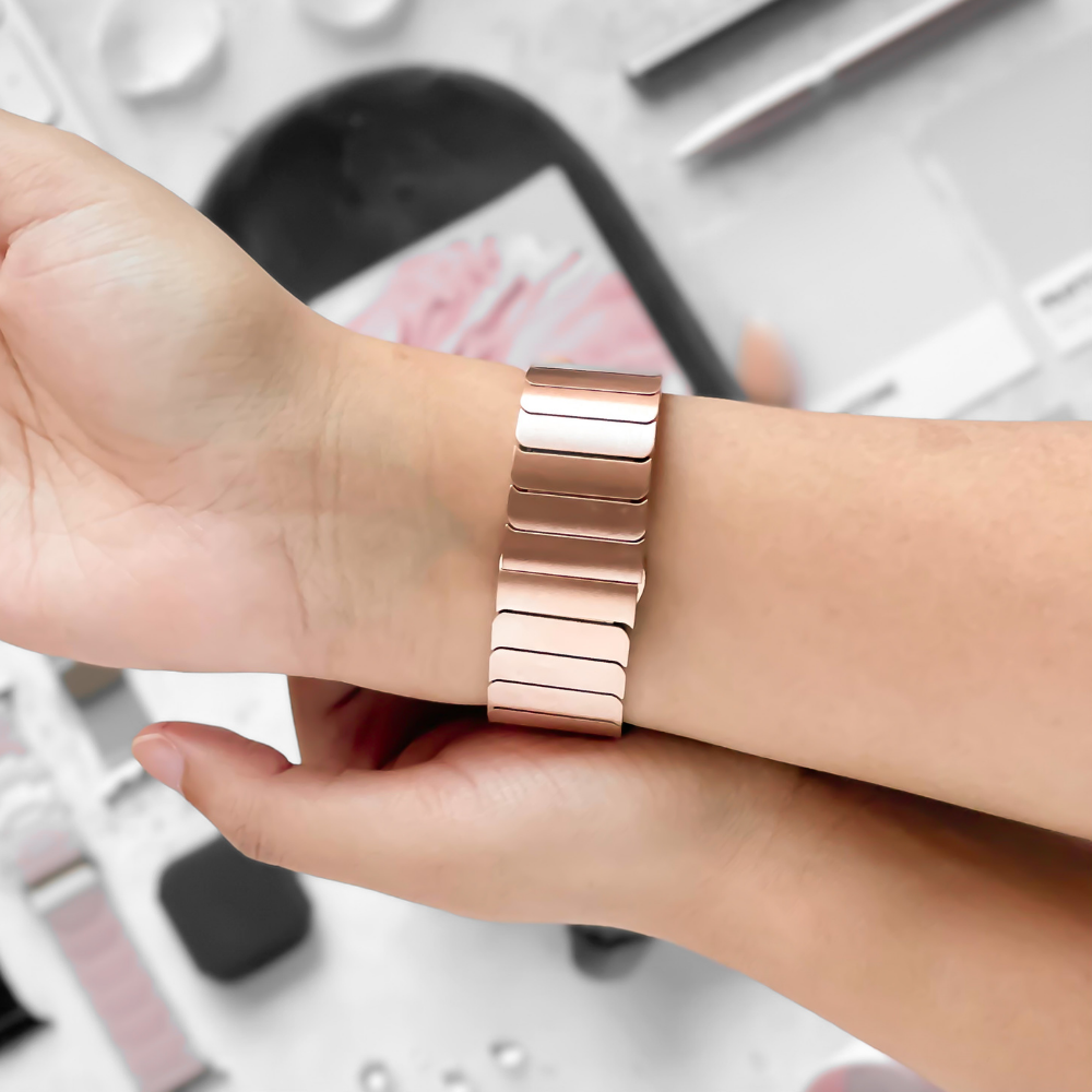 A person adjusts a Coconut Lane Slinky Apple Watch Strap in rose gold on their wrist. The smartwatch's rectangular links create a sleek and modern design. The background is blurred, featuring various smartwatch accessories and a smartphone.