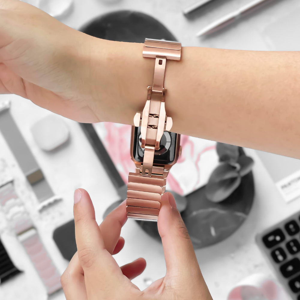A person fastening a rose gold Slinky Apple Watch Strap from Coconut Lane onto their wrist. The background features various items, including bands and tools, in a blurred setting.