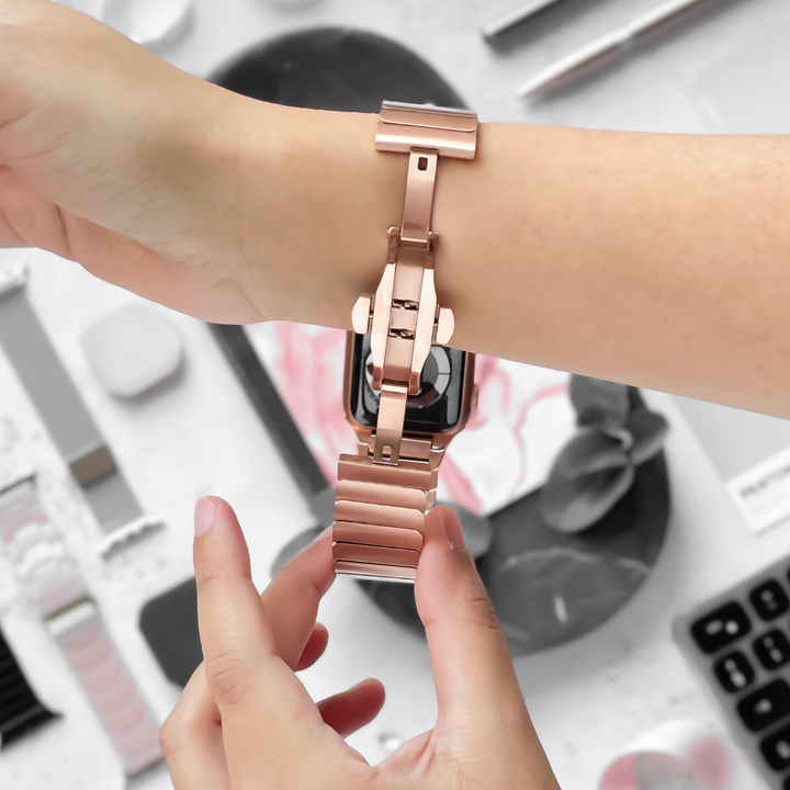 A person fastening a rose gold Slinky Apple Watch Strap from Coconut Lane onto their wrist. The background features various items, including bands and tools, in a blurred setting.
