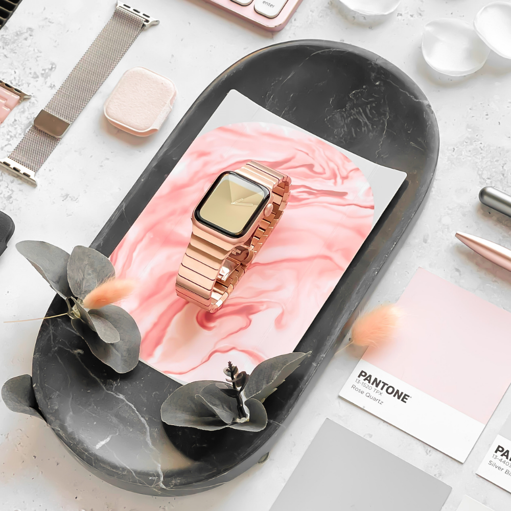 The Slinky Apple Watch Strap - Rose Gold from Coconut Lane is elegantly displayed on a black marble tray, accompanied by other accessories such as a Zinc Alloy Adjustable strap, a pink makeup compact, eucalyptus leaves, and scattered Pantone color swatches on the marble surface.