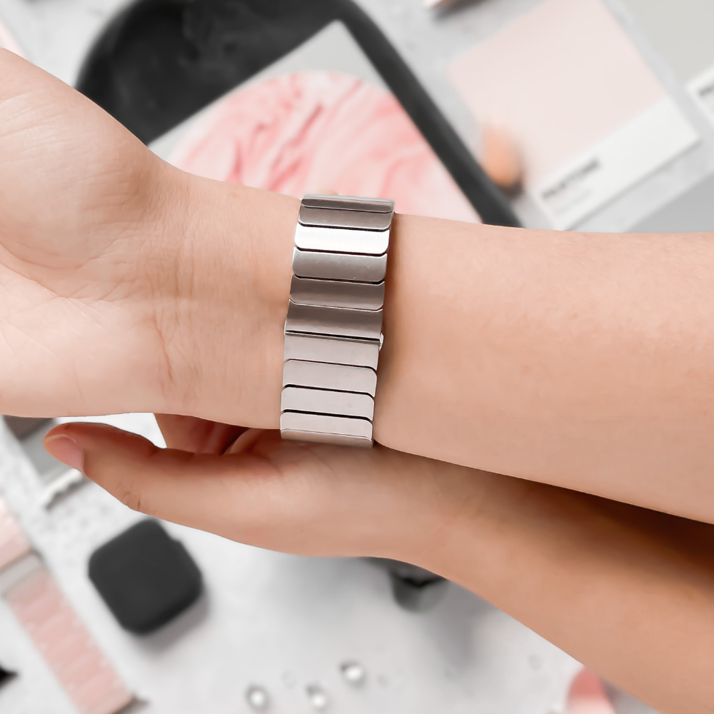 A close-up of a person adjusting a sleek Coconut Lane Slinky Apple Watch Strap in Silver on their wrist. The background features various pink and grey swatches, creating a stylish and aesthetically pleasing scene. An adjustable strap ensures the perfect fit, with a FREE link remover tool for easy customization.
