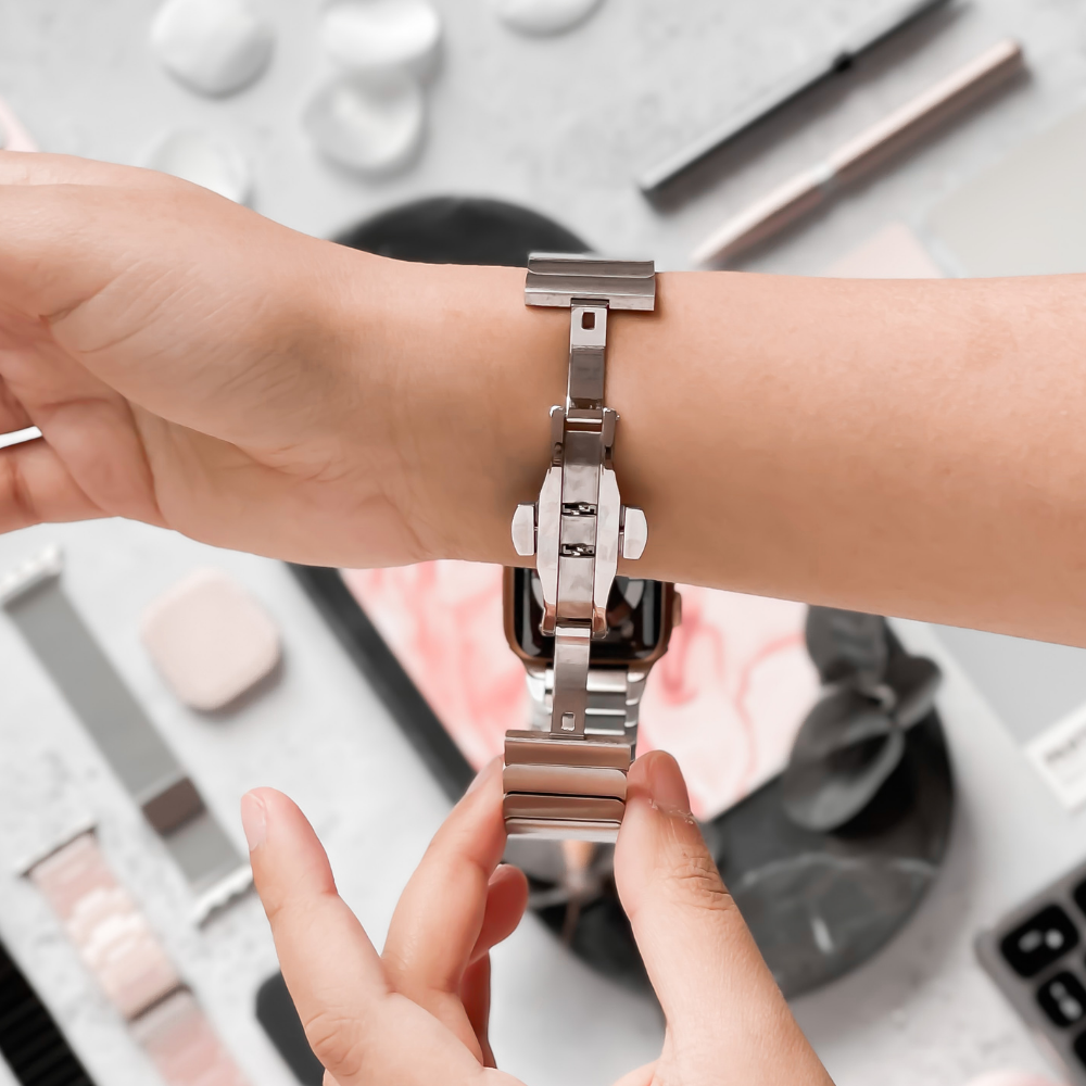 A person is adjusting a Coconut Lane Slinky Apple Watch Strap - Silver on their wrist. The background features an array of beauty and grooming items, including makeup palettes, brushes, and skincare products, all neatly arranged on a flat surface. The focus is on the adjustable strap and the person's hands using a FREE link remover tool.