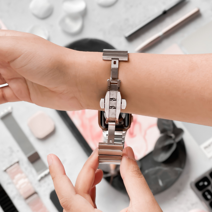 A person is adjusting a Coconut Lane Slinky Apple Watch Strap - Silver on their wrist. The background features an array of beauty and grooming items, including makeup palettes, brushes, and skincare products, all neatly arranged on a flat surface. The focus is on the adjustable strap and the person's hands using a FREE link remover tool.