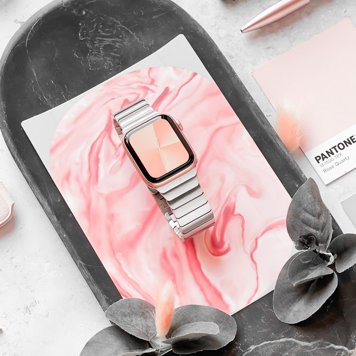 The Coconut Lane Slinky Apple Watch Strap - Silver, featuring a chic silver metal band, is elegantly displayed on a pink and white marble-patterned surface. Nearby, green leaves and a pale pink Pantone card contribute to an aesthetically pleasing and harmonious flat lay composition. The package also includes a FREE link remover tool for the adjustable strap.