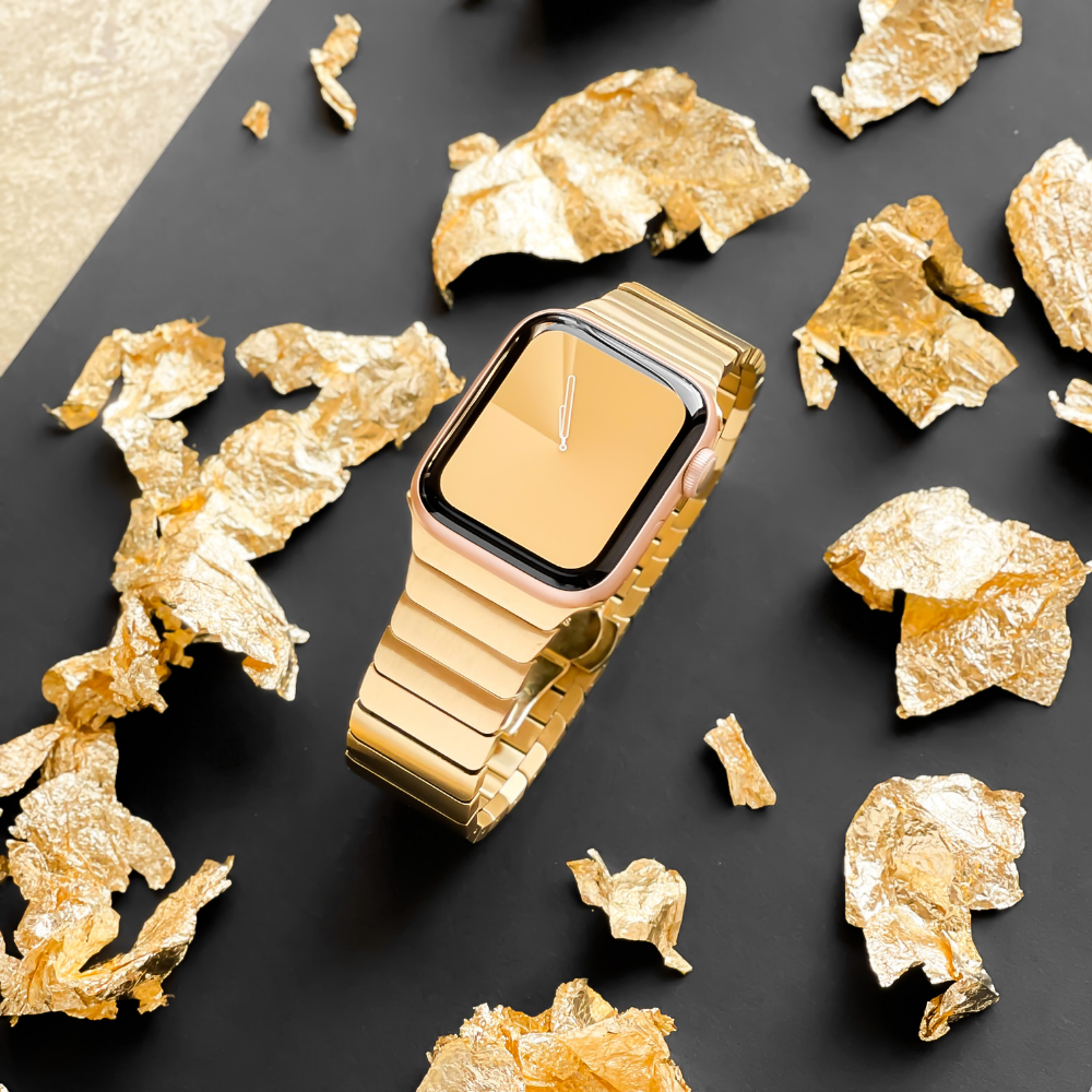 A gold smartwatch with a matching metal band is displayed on a black surface. Surrounding the watch are scattered pieces of gold foil, contributing to a luxurious and elegant aesthetic, accentuated by the sophistication of the Slinky Apple Watch Strap - Gold from Coconut Lane.