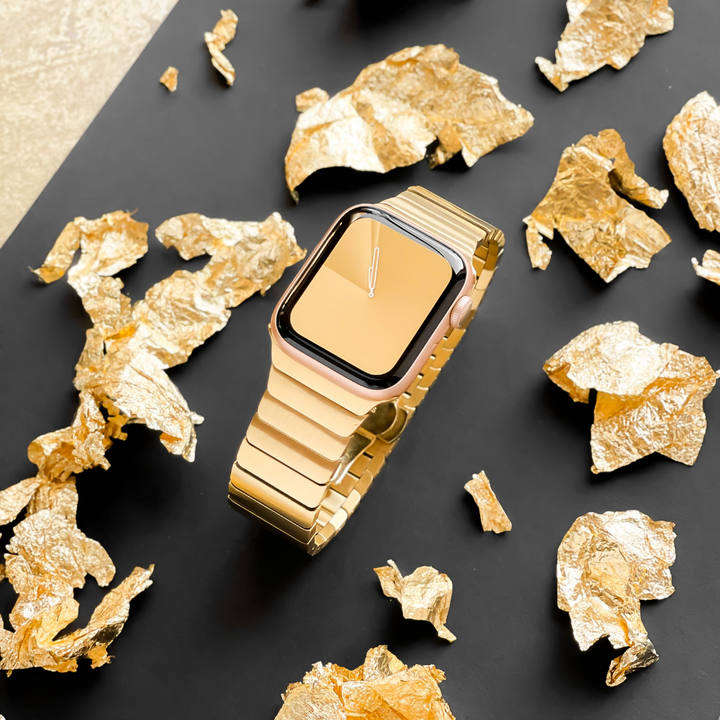 A gold smartwatch with a matching metal band is displayed on a black surface. Surrounding the watch are scattered pieces of gold foil, contributing to a luxurious and elegant aesthetic, accentuated by the sophistication of the Slinky Apple Watch Strap - Gold from Coconut Lane.