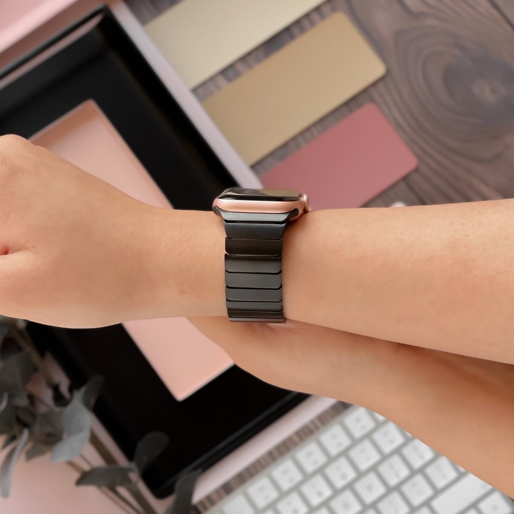 A close-up of a person's wrist wearing an Apple Watch with the Coconut Lane Slinky Apple Watch Strap in Black, featuring a dark metallic link band. The background includes a keyboard, some decorative items, and color swatches in shades of pink, beige, and brown.