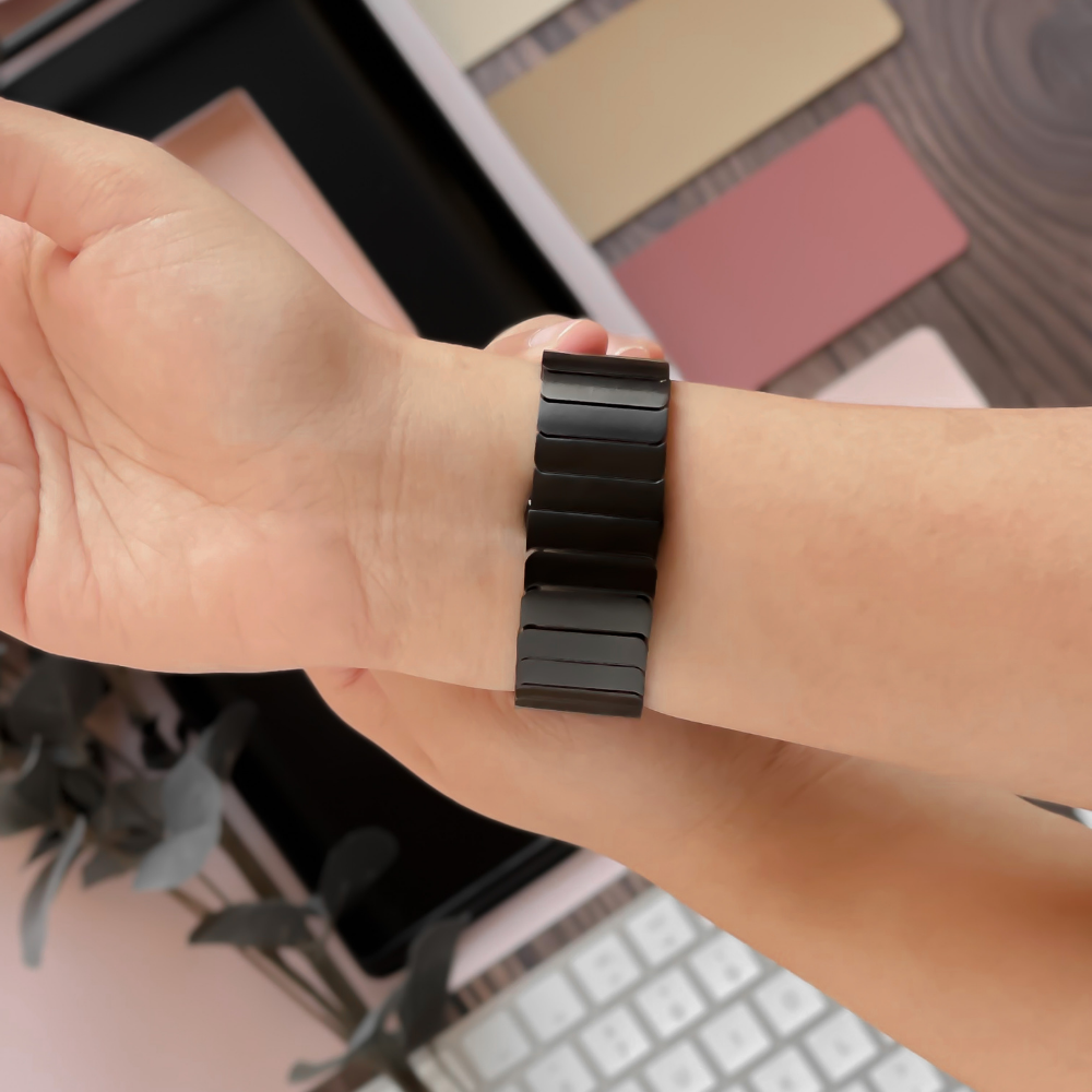 A person's wrist is shown wearing a sleek Coconut Lane Slinky Apple Watch Strap - Black. The background features pastel-colored cards, a white keyboard, and a plant with dark green leaves. The image exudes a soft and bright aesthetic, making the adjustable Zinc Alloy strap stand out effortlessly.