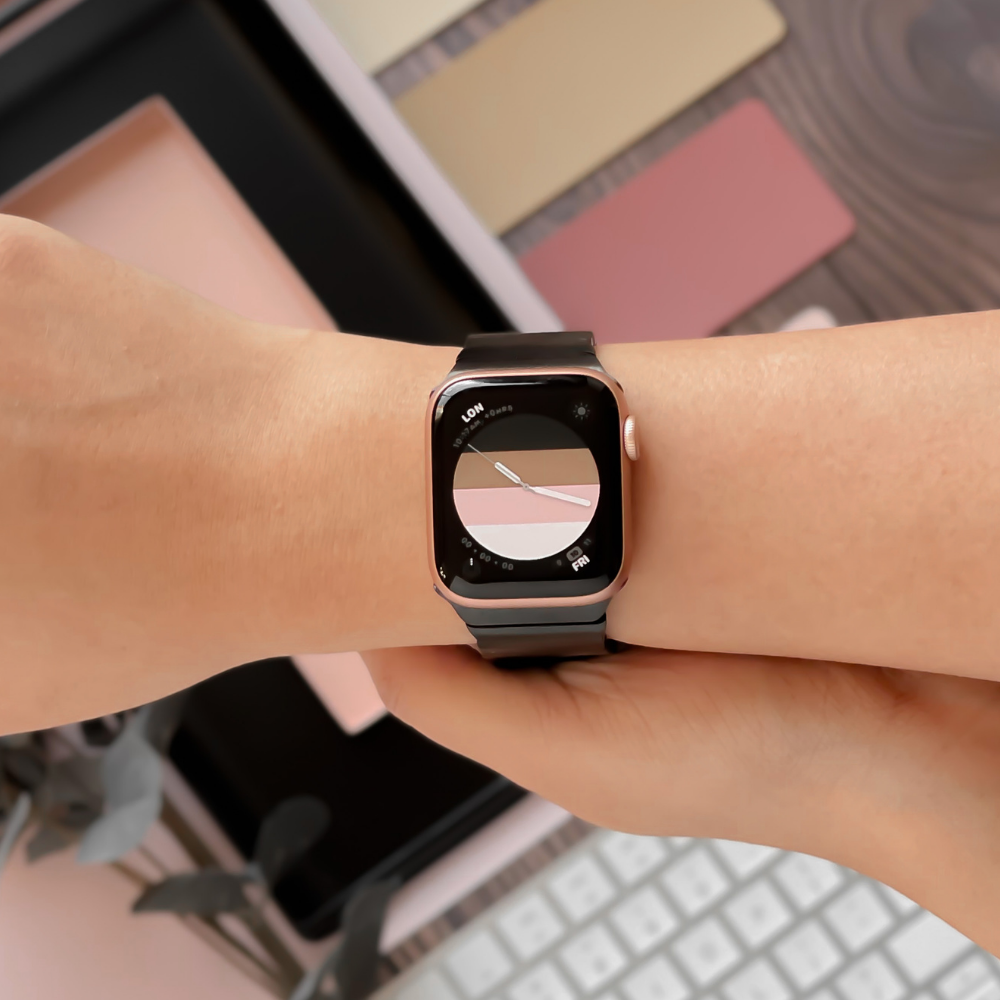 A person wearing a smartwatch featuring a modern, minimalist watch face and a sleek Slinky Apple Watch Strap - Black by Coconut Lane. The background contains various objects such as a keyboard, swatches of pastel-colored materials, and a plant, creating a creative and stylish workspace vibe that complements all Apple Watch models.