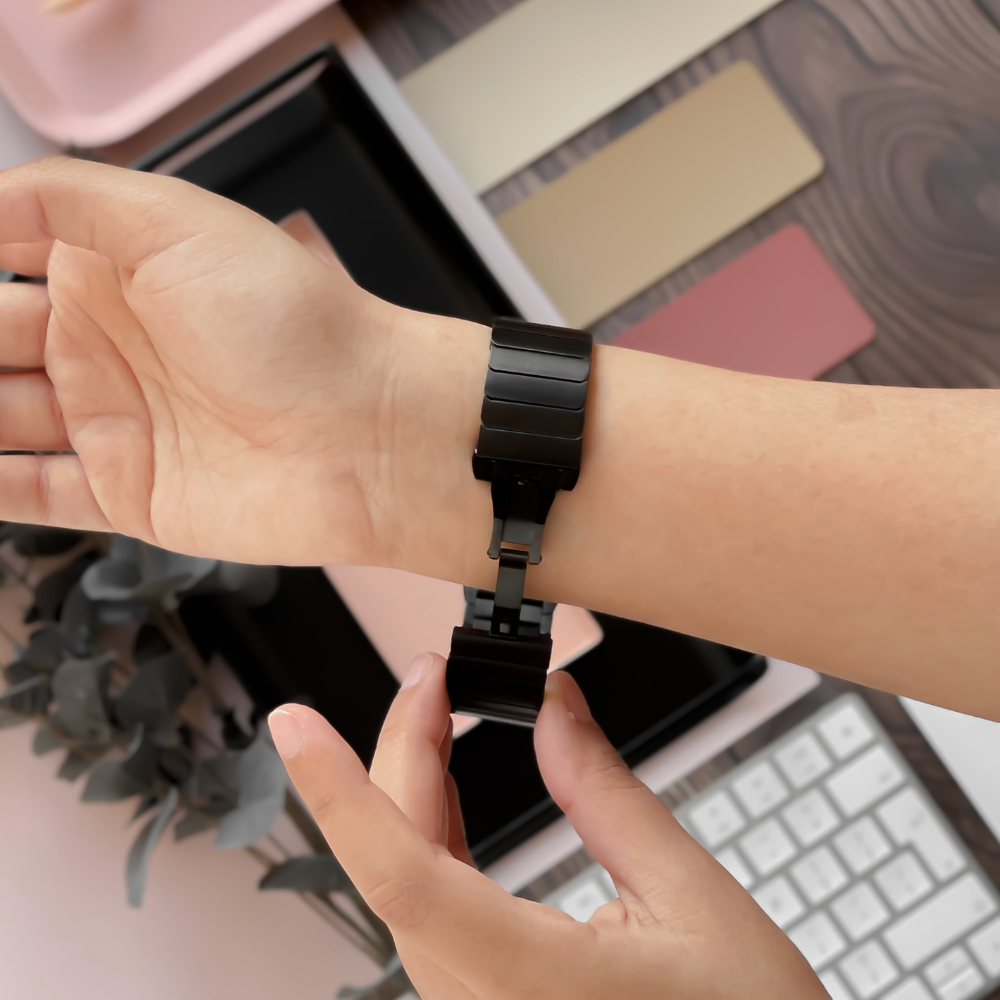 A person is adjusting the clasp of a Coconut Lane Slinky Apple Watch Strap - Black on their wrist. A blurred computer keyboard, color swatches, and some eucalyptus leaves are visible in the background. The setting appears to be a workspace or desk.