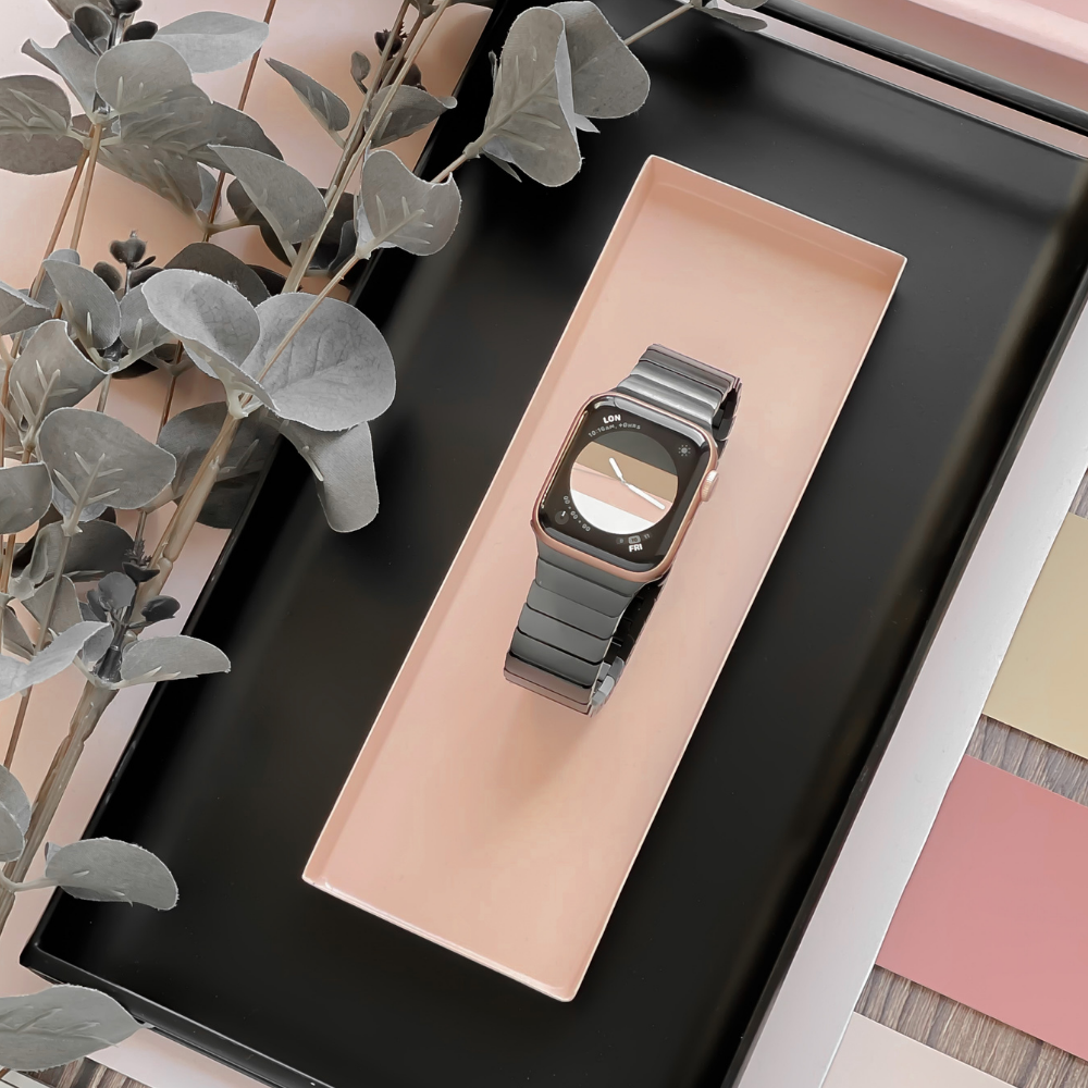 A Slinky Apple Watch Strap - Black by Coconut Lane is displayed on a rectangular tray with a pastel pink base. The tray is set on a larger black tray, surrounded by eucalyptus leaves and color swatches in soft shades on a wooden surface.