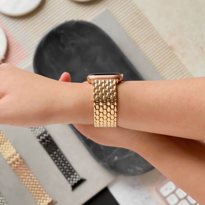 A person is wearing a Mermaid Apple Watch Strap - Gold by Coconut Lane on their wrist. Various other smartwatch straps, including black and patterned options, along with a zinc alloy adjustable strap, are displayed on a nearby table. The background features a mix of textured surfaces.