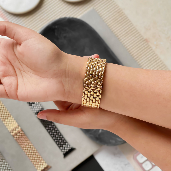 A person trying on the wide, gold, Mermaid Apple Watch Strap from Coconut Lane on their wrist. The background features various other bracelet options, including Zinc Alloy Adjustable Straps in different colors and patterns, laid out on a textured surface.
