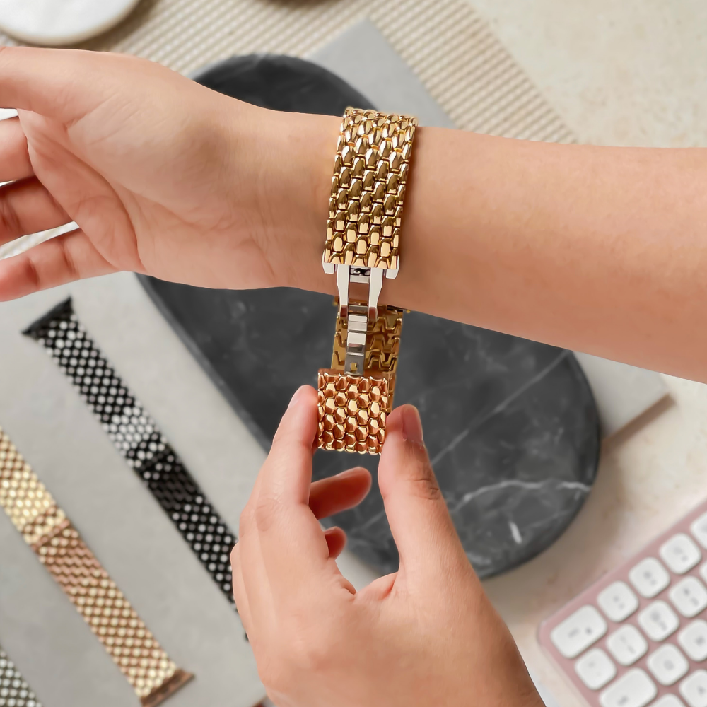 A close-up of hands adjusting a Coconut Lane Mermaid Apple Watch Strap - Gold. The band is being fastened onto a wrist. Black, white, and gold watch bands are laid out on a textured surface in the background, alongside a pink and white keyboard partially visible on the right.