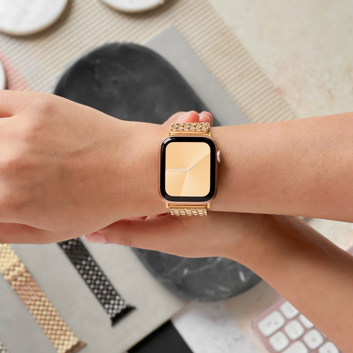 A person is wearing a gold-colored Apple Watch with the Coconut Lane Mermaid Apple Watch Strap - Gold, which features a zinc alloy adjustable strap. The watch face displays a simplistic clock with white hour and minute hands on a light yellow background. Below on the table, various other watch bands in different styles and colors, along with a link remover tool, are visible.