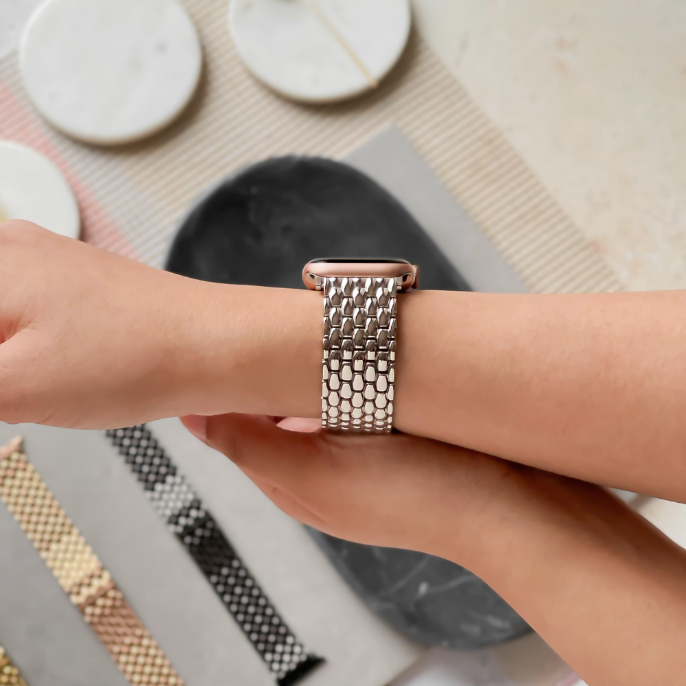 A person with light skin is adjusting a Coconut Lane Mermaid Watch Strap - Silver on their wrist. The watch features a metallic mesh band. Various other watch bands, coasters, and a link remover tool are scattered on a textured surface in the background.