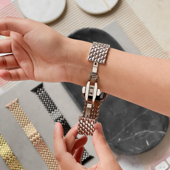 A person fastens a metallic mesh watch band around their wrist. In the background, a variety of other watch bands lie on a textured surface, including the Coconut Lane Mermaid Watch Strap - Silver, available in stunning shades such as gold, black, and silver.