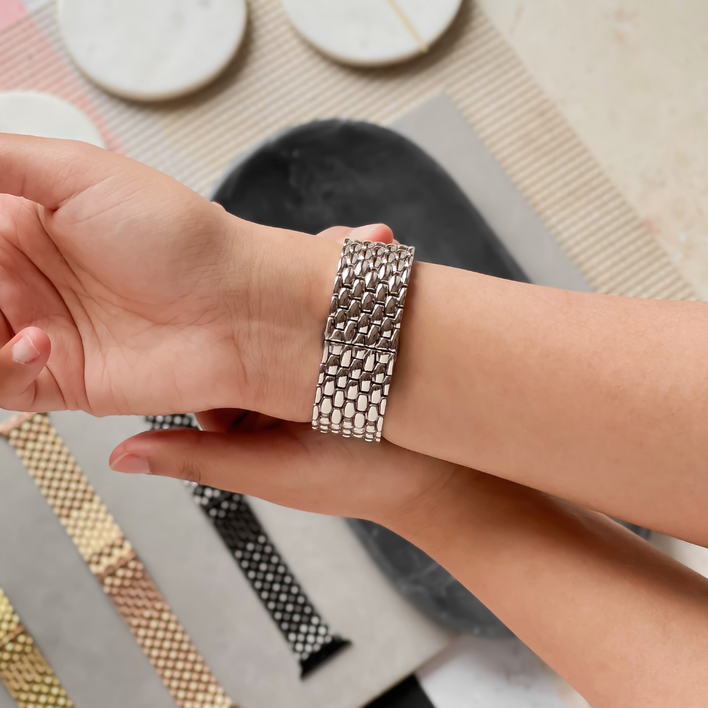 A person’s wrist adorned with Coconut Lane's Mermaid Watch Strap - Silver. The background displays an array of watch bands in diverse colors and patterns, neatly arranged on a table, including the Silver Mermaid Watch Strap.